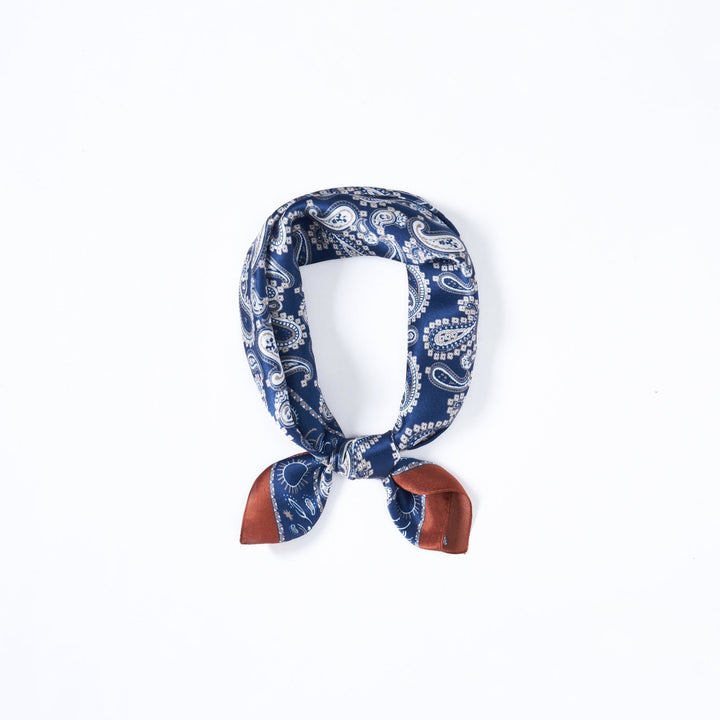 A Paisley Series Square Silk Scarf by SusanSilk, featuring blue and white paisley patterns with brown tips, is tied into a loop on a white background.