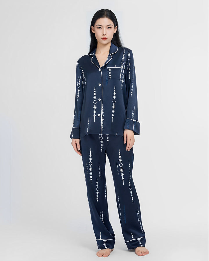 A woman is standing barefoot in the Diamond Striped Silk Pajamas Set by SusanSilk, featuring a long-sleeve, buttoned design in navy blue with a white geometric pattern. The background is plain white.