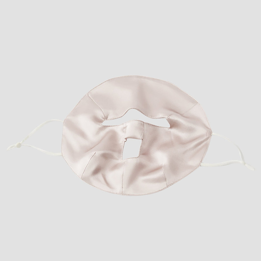 The Beauty Silk Face Mask by SusanSilk is a beige silk face mask with a unique design featuring multiple cutouts, including a square cutout for the mouth area and a horizontal slit for the nose. It has white elastic ear loops, effortlessly combining style and comfort.