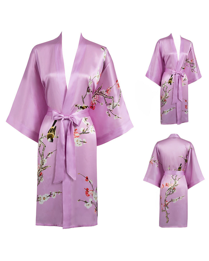 The Plum Blossom Short Silk Kimono by SusanSilk is a light purple satin robe adorned with intricate floral and bird embroidery. This luxurious silk kimono features wide sleeves, a waist tie, and a knee-length cut. Three views of the kimono are shown: front, side, and back.