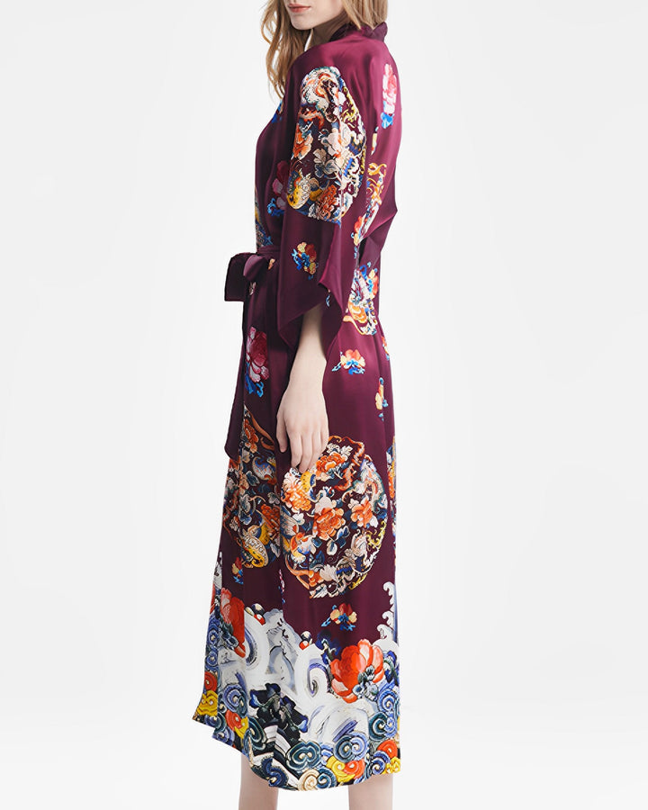 A person wearing the Wheel of Fortune Long Silk Kimono by SusanSilk, featuring Japanese-inspired designs in a luxurious burgundy pattern, stands facing slightly to the side.#color_red-wine