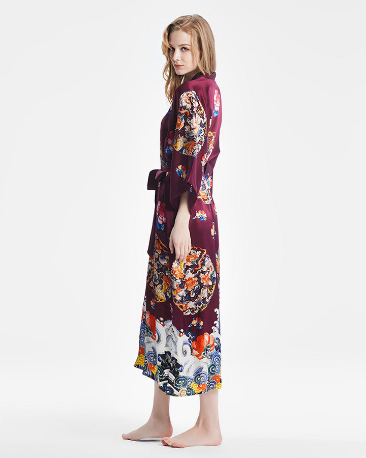 A person stands barefoot, wrapped in a Wheel of Fortune Long Silk Kimono by SusanSilk, looking over their shoulder against a plain white background.#color_red-wine