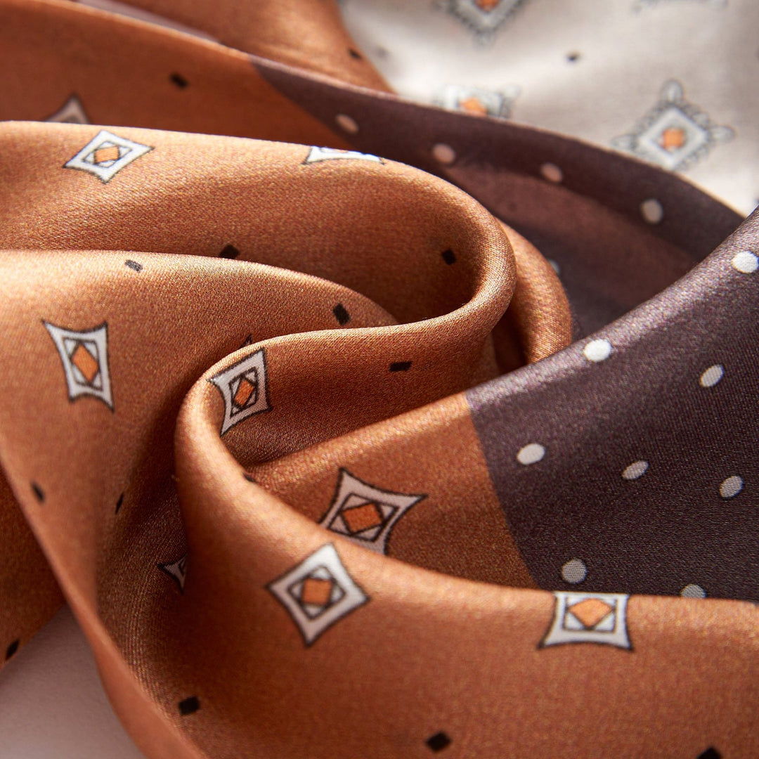 Close-up of the SusanSilk Tri-Colored Gemstone Narrow Long Silk Scarf, showcasing a geometric pattern in shades of brown and beige with small diamond shapes and dots. The fabric appears smooth and lustrous, with gentle folds and creases.